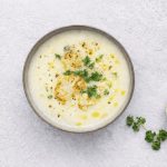 Zuppa di cavolfiore: leggera, calda e facile da preparare 1 In primo piano su un tavolo chiaro c'è una zuppa di cavolfiore condita con olio EVO e del prezzemolo riccio.