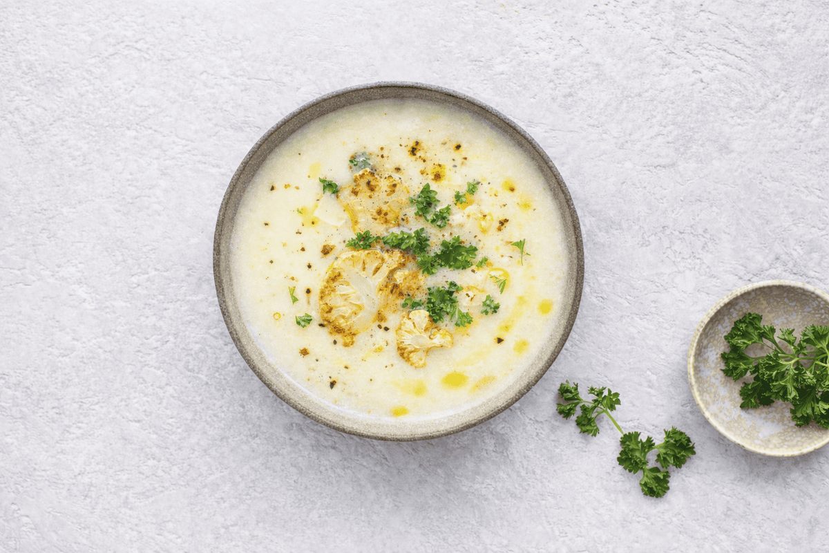 In primo piano su un tavolo chiaro c'&egrave; una zuppa di cavolfiore condita con olio EVO e del prezzemolo riccio.