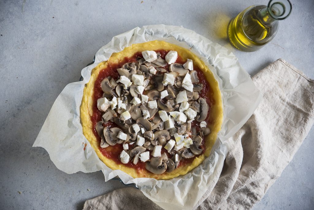 Pizza di polenta con pomodoro, mozzarella e funghi 7 un teglia tonda su panno bianco con dentro la ricetta con polenta: pizza di polenta