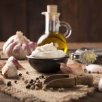 Salsa aioli: ricetta sana e leggera con aglio 1 su tavolo di legno, sottopiatto in Juta con ciotola di terracotta con la salsa aioli e ingredienti come aglio, olio d'oliva e pepe