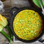Frittata di zucchine, un classico gustoso, veloce e completo 2 su base di legno una padella tonda con i manici e dentro una frittata di zucchine, accanto due zucchine col fiore