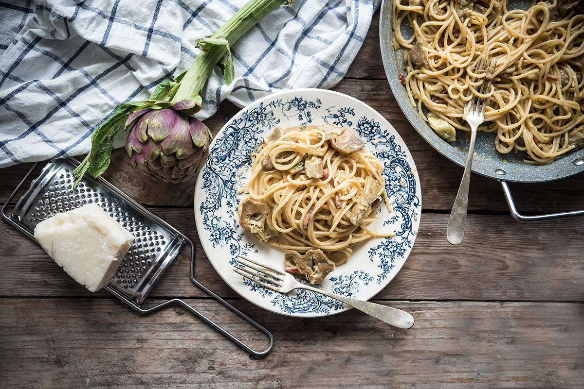 su base di legno un piatto decorato con spaghetti e una forchetta, accanto una padella con spaghetti alla carbonara di carciofi