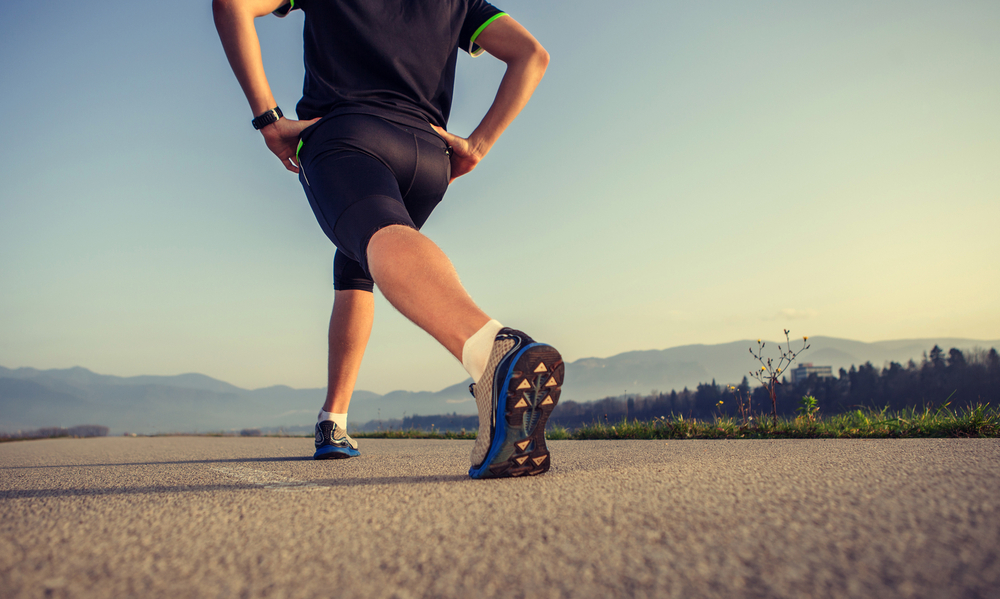 Riscaldamento muscolare: che cos'è, i benefici e gli esercizi da fare 8 In primo piano un atleta con un completo sportivo nero mentre esegue il riscaldamento muscolare prima di una corsa su strada asfaltata.