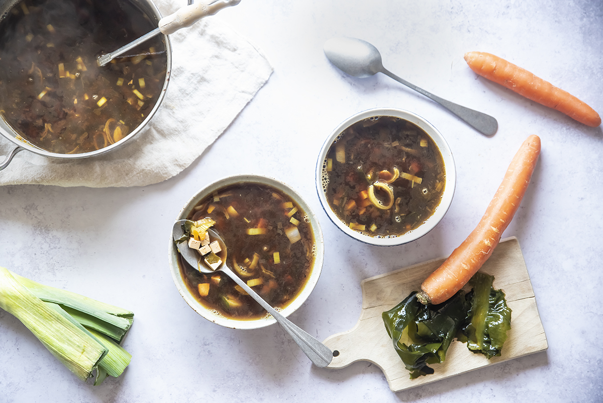 Minestre e zuppe: ricette da gustare quando fa freddo e non solo 19 su tavolo di marmo 2 contenitori bianchi con la zuppa di miso e attorno gli ingredienti: carota ,1 porro 2 cucchiai, e dietro la zuppiera piena di zuppa