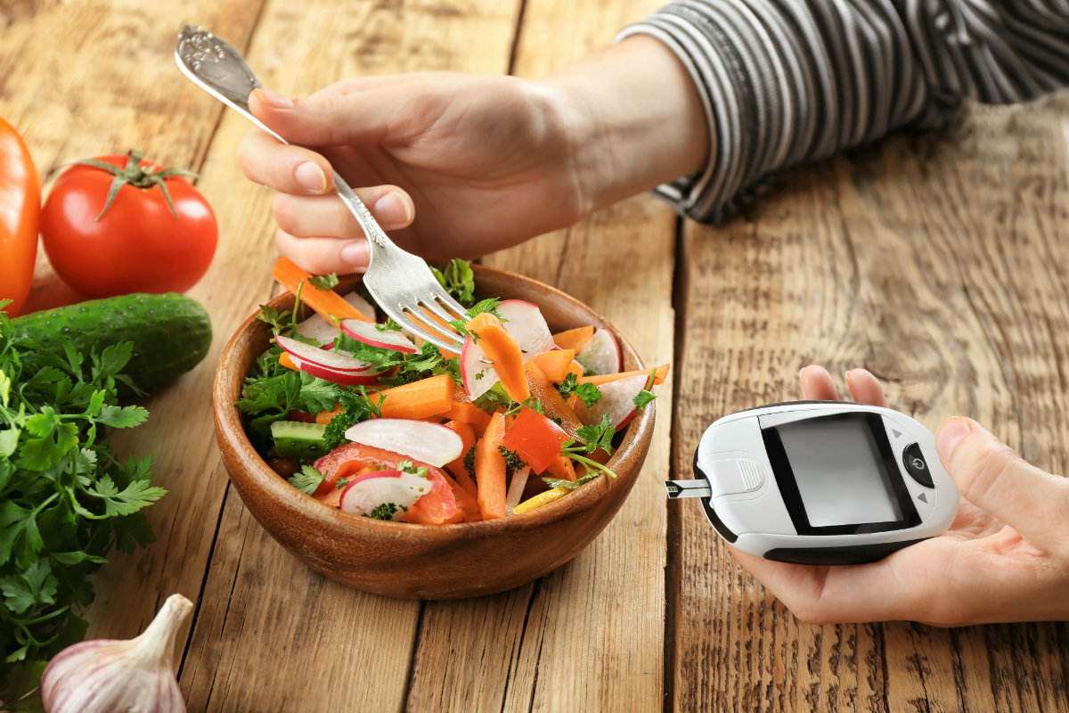 Dieta per il diabete: cosa mangiare e esempio di menù settimanale 1 su tavolo di legno, si vede mani du una persona con la forchetta che mangia un insalata e che tiene nell'altra mano un glucometro per sorvegliare la sua dieta per il diabete