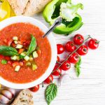 Gazpacho: ricetta e video ricetta, storia e varianti light 1 su tavolo di legno bianco, una scodella con dentro il gazpacho, attorno pomodorini, peperone verde limone e una bottiglia di olio