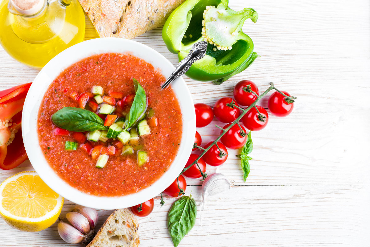 su tavolo di legno bianco, una scodella con dentro il gazpacho, attorno pomodorini, peperone verde limone e una bottiglia di olio
