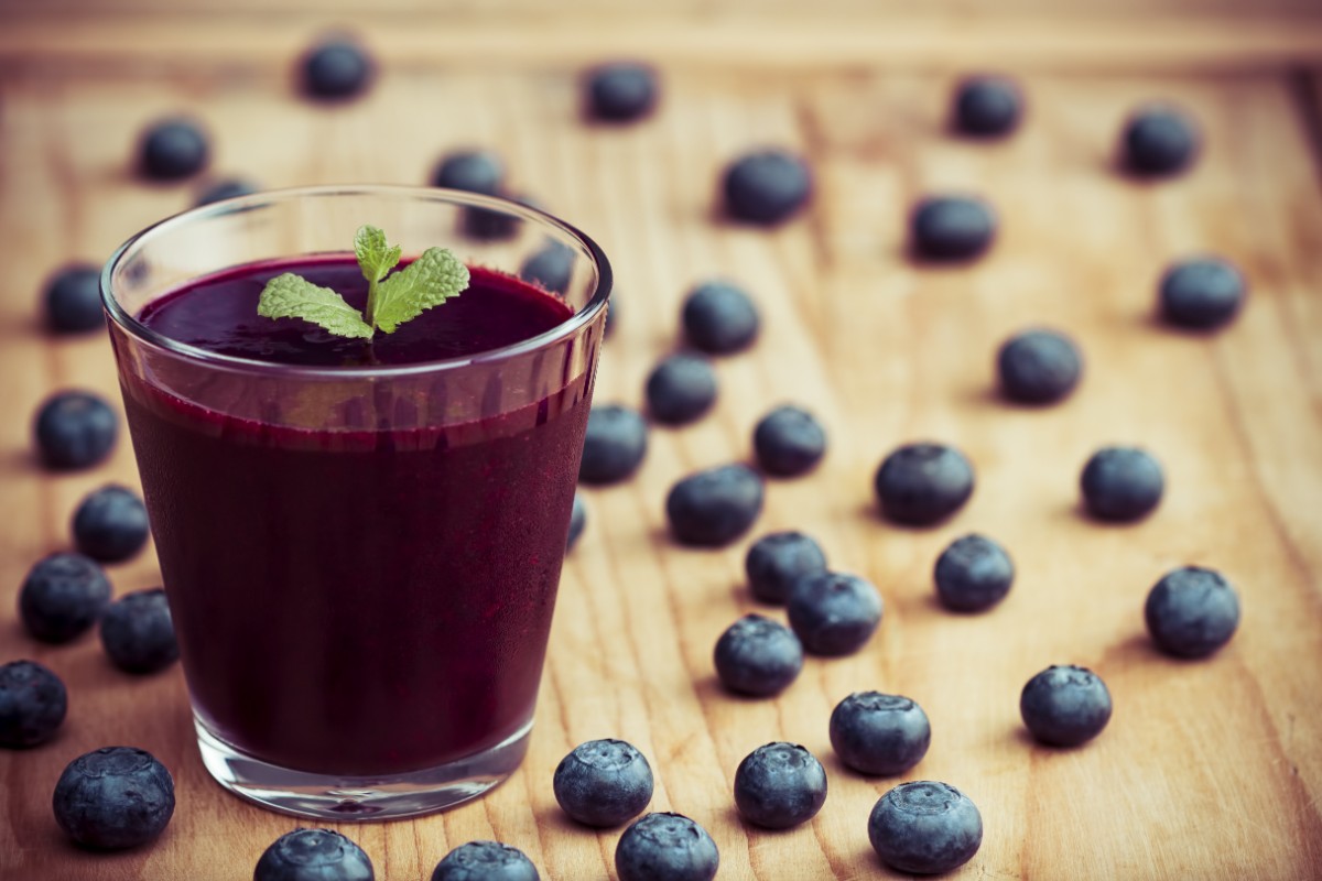 Mirtilli: proprietà, calorie, controindicazioni e ricette 2 Un bicchiere colmo di succo di mirtillo e alcuni mirtilli sparsi su un tavolo