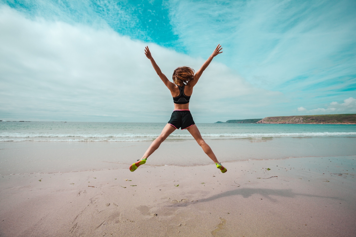 Kayla Itsines e l'allenamento bikini workout: che cos'è, benefici, video allenamento 1 una giovane donna in bikini salta a braccia aperte al centro di una spiaggia deserta