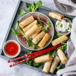 Involtini primavera al forno: la ricetta leggera 1 vassoio rettangolare con tanti involtini primavera, alcuni in un piatto tondo, ciotola salsa agrodolce e bacchette cinesi
