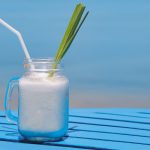 Virgin Colada, il cocktail analcolico esotico 1 con sfondo di mare, tavolo azzurro con bicchiere con manico con un virgin colada, foglia di ananas e cannuccia