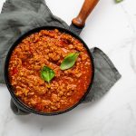Il ragù alla bolognese: una ricetta leggera ma ricca di gusto! 1 In primo piano su uno sfondo di marmo chiaro una pentola nera con il ragù alla bolognese, decorato con una foglia di basilico.
