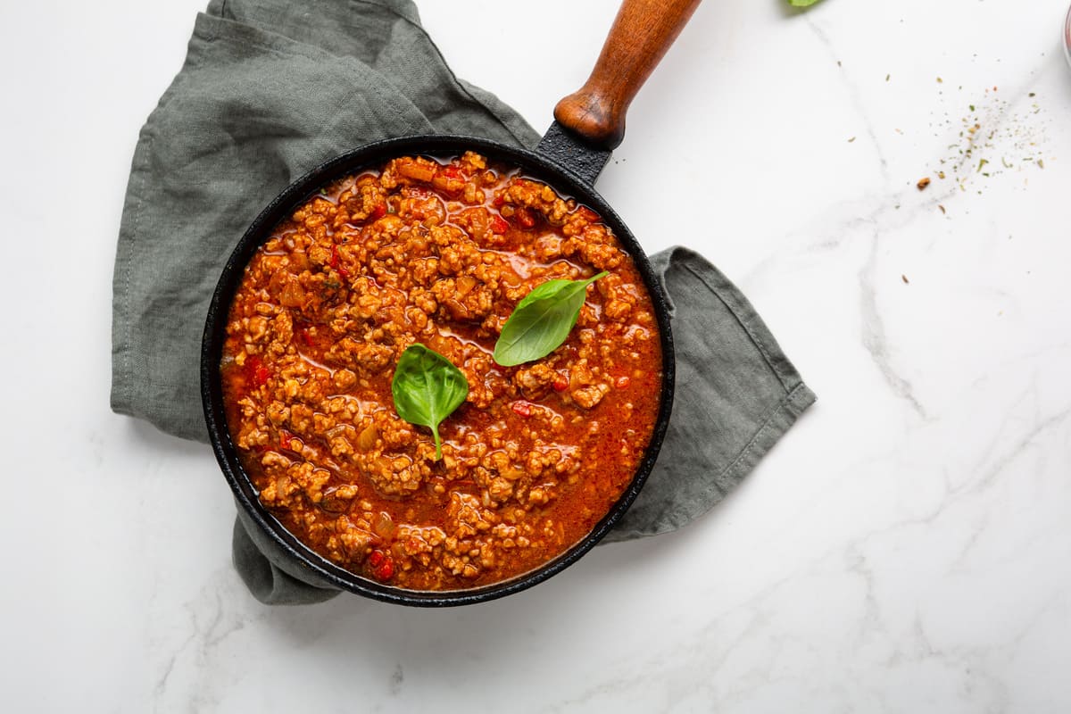 In primo piano su uno sfondo di marmo chiaro una pentola nera con il rag&ugrave; alla bolognese, decorato con una foglia di basilico.