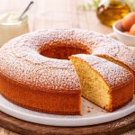 Torta 7 vasetti: la ricetta facile e soffice per la colazione 1 In primo piano su un tavolo di legno rustico c'è un piatto fondo con la torta 7 vasetti. Sullo sfondo un vasetto di yogurt, olio EVO e delle uova.