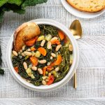 Zuppa di cavolo nero e fagioli: ricetta facile e cremosa 1 In primo piano su un tavolo di legno chiaro, una zuppa di cavolo nero con fagioli, carote e una fetta di pane tostato. Sulla destra un cucchiaio d'oro satinato. In alto a sinistra delle foglie di cavolo nero.