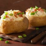 Patate ripiene al forno: la ricetta per farle croccanti fuori e morbide dentro 1 su tavolo di legno, tagliere di legno con 2 patate ripiene al forno, accanto forchetta e coltello