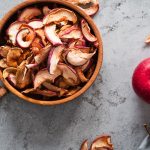 Mele essiccate: ricetta facile per uno snack croccante 1 In primo piano su un tavolo di legno c'è una ciotola con delle mele essiccate, accanto dei spicchi di mela essiccati e una mela rossa.