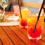 Spritz analcolico: la ricetta con arancia e bitter analcolico 1 su tavolo di legno, 2 bicchieri con spritz analcolico, ghiaccio, limone e cannucce accanto un tagliere di legno con olive, pizette e tramezzini, accanto busto di una dona con maglietta nera che sta bere aperitivo