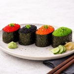 Gunkanmaki con uova di lompo: ricetta facile con foto passo passo 1 In primo piano su un tavolo chiaro c'è un piatto con dei gunkanmaki di lompo, delle fettine di cetriolo, zenzero e salsa wasabi. Sulla destra su un tovagliolo grigio ci sono due bacchette in legno.