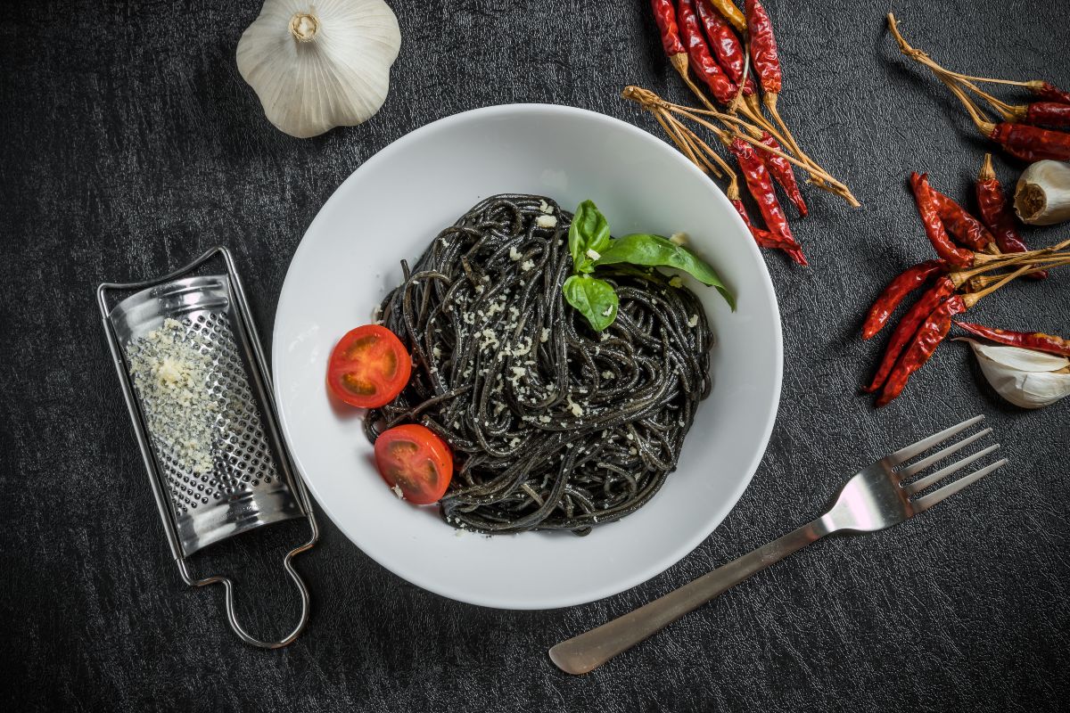 Spaghetti al nero di seppia: un primo piatto siciliano 2 su un tavolo scuro, primo piano di un piatto bianco con gli spaghetti al nero di seppia e attorno gli ingredienti come peperoncini e aglio.