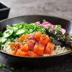 Poke bowl: ingredienti, ricetta e video ricette 2 su tavolo di marmo: ciotola con poke bowl con alghe e salmone. Accanto bacchette cinese e dietro ciottola piccola con salsa di soia