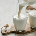 Yogurt fatto in casa, una ricetta semplice con solo 2 ingredienti 1 su tavolo di legno bianco, tagliere di legno con 2 bicchieri con yogurt fatto in casa e una mano che versa il latte sopra