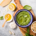 Pesto alla genovese: la ricetta originale per farlo in casa 1 su piano di lavoro di marmo tagliere con una ciotola piena di pesto alla Genovese e attorno gli ingredienti, limone, basilico, aglio e basilico. Sul tagliere ciotola piena di pinoli
