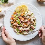 Insalata di polpo, la ricetta step by step 1 su tavolo bianco, canovaccio beige con piatto bianco con 2 mani in campo . La mano destra tiene la forchetta e sta per prendere un pò di insalata di polpo con carote e sedano. Attorno limone tagliato in 2.