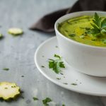 Vellutata di zucchine, un primo leggero e facile da preparare 1 su base grigia piattino e tazza da brodo bianca piena di vellutata di zucchine. Rondelle di zucchine sparse