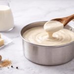 Besciamella fatta in casa: ricetta facile pronta in 5 minuti 1 In primo piano su un tavolo chiaro c'è un pentola con la besciamella, sullo sfondo gli ingredienti della ricetta come noce moscata, burro e latte.