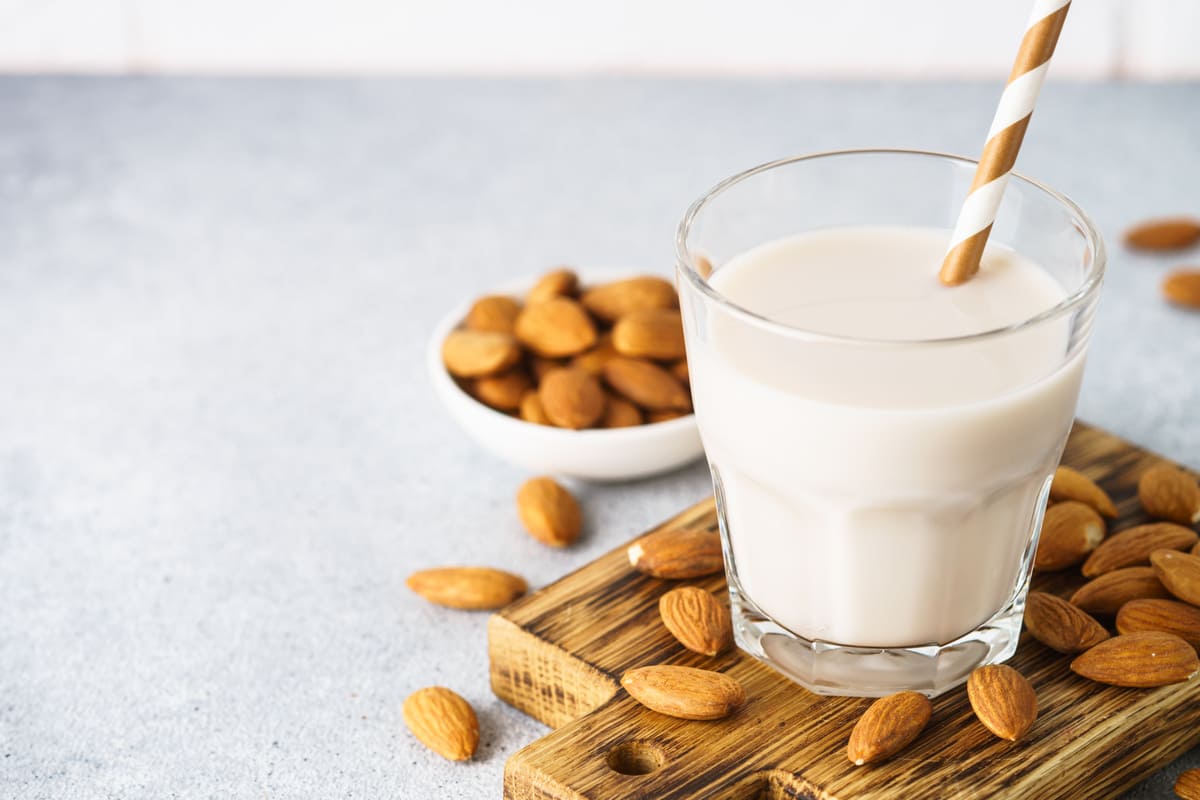 Ricette con le mandorle: tante idee gustose da provare 34 In primo piano su uno sfondo chiaro c'è un tagliere di legno con un bicchiere di latte di mandorla e una cannuccia. Sullo sfondo ci sono delle mandorle.