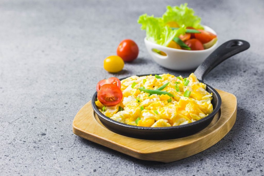 Colazione alternativa: 8 idee per una colazione sana 5 su vassoio di legno una padella con manico piena di uova strappate e un pomodoro a metà