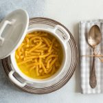 Passatelli in brodo, prepara il primo della tradizione romagnola 1 su tavolo di marmo . piatto di argento con una zuppiera che contiene i passatelli in brodo . Sulla destra un tovagliolo di cottone con un cucchiaio legato con un filo con carta