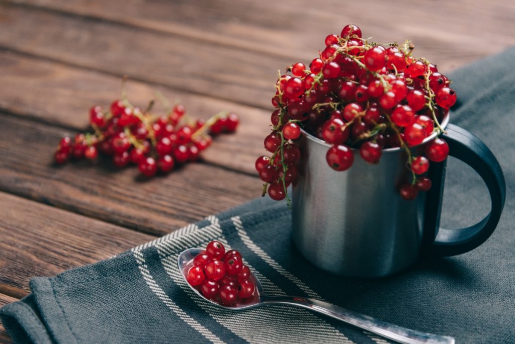 Mirtillo rosso o cranberry: il superfood tra proprietà, benefici, ricette e i migliori integratori 2 su tavolo di legno, tovagliolo con tazza piena di mirtillo rosso americano, accanto cucchiaino con bacche di frutti rossi