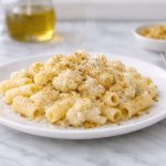 Pasta con cavolfiore: primo piatto leggero e cremoso 1 In primo piano su un tavolo di marmo chiaro c'è un piatto di pasta con cavolfiore con del formaggio e noce moscata. Sulla destra c'è un tovagliolo grigio con una forchetta.