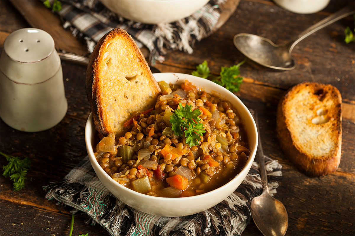 ricette con lenticchie: su tavolo di legno una zuppiera con zuppa di lenticchie e un crostino di pane, saliera e altro pane accanto
