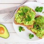 Avocado toast: una ricetta sana e leggera 1 su base di legno chiaro mezzo avocado e un piatto bianco con due avocado toast