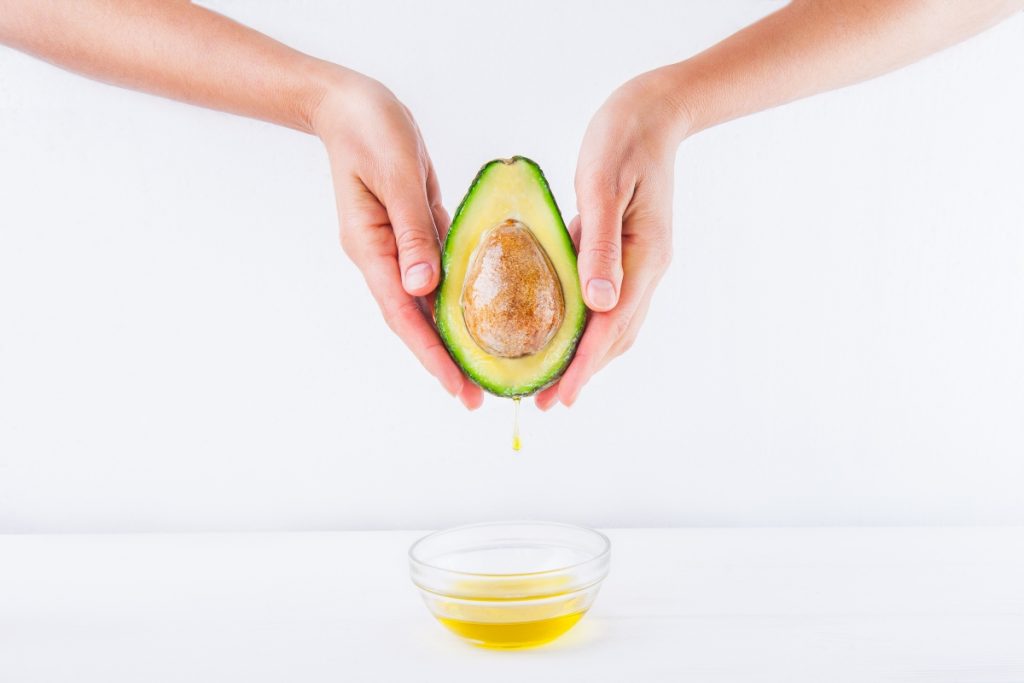 Olio di avocado: proprietà, usi e come farlo in casa 4 due mani femminili in primo piano che tengono in mano mezzo avocado e sotto una ciotolina con dell'olio di avocado