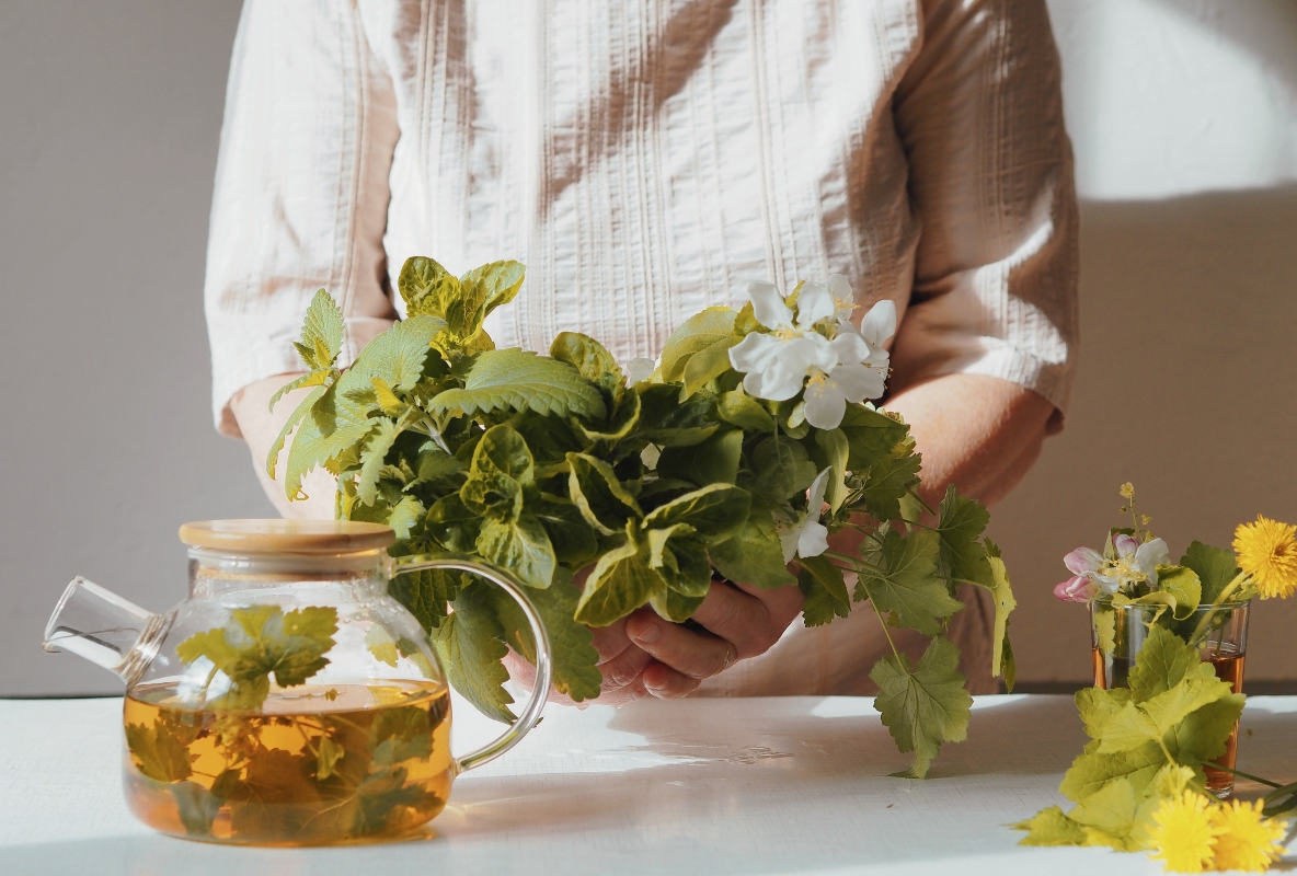 donna in camicia bianca davanti al bancone della cucina che sta preparando uno dei rimedi naturali anticellulite con delle piante per farsi una tisana
