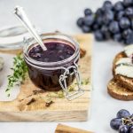 Marmellata di uva nera fatta in casa: la ricetta senza zucchero da provare 2 In primo piano sopra una tavoletta di legno, c'è un barattolo di vetro con la marmellata di uva nera, fette di pane con formaggio e chicchi d'uva.