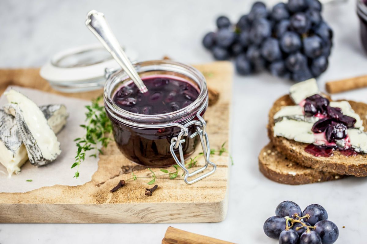 Ricette con uva: 12 idee leggere e gustose 12 In primo piano sopra una tavoletta di legno, c'è un barattolo di vetro con la marmellata di uva nera, fette di pane con formaggio e chicchi d'uva.