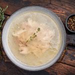 Brodo di pollo fatto in casa: ricetta e video ricetta passo passo 1 In primo piano in un piatto fondo e su un tavolo di legno scuro, brodo di pollo con erbe aromatiche e pepe nero ai lati.