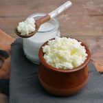Kefir di latte, come preparare in casa la bevanda sana e buona 1 su tavolo da cucina tagliere di legno con un contenitore di kefir di latte e un barattolo con latte e un cucchiaino di legno