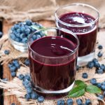 Succo di mirtillo senza zucchero: tutti i benefici e come prepararlo in casa 2 su tavolo di legno 2 bicchieri con succo di mirtillo e attorno mirtilli freschi