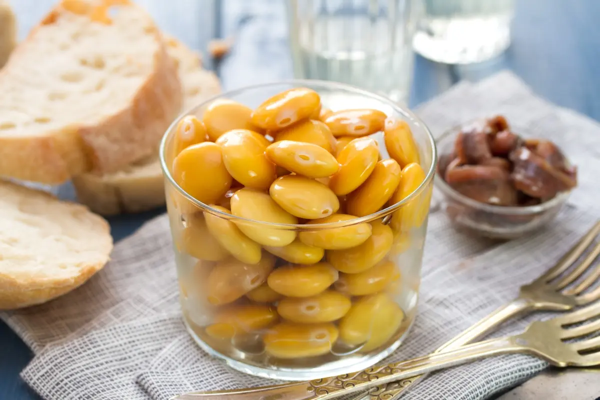 Lupini: calorie, proprietà e come mangiarli 1 su tavolo primo piano di un bicchiere pieno di lupini con 2 forchette e accanto del pane e dietro un bicchiere d'acqua