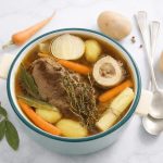 Brodo di carne fatto in casa: ricetta base e consigli di cottura 1 In primo piano su un tavolo di marmo chiaro c'è una pentola in ceramica bianca con del brodo di carne. Intorno c'è una foglia di allora, una carota, una patata e delle posate.