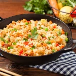 Riso alla cantonese: ricetta e video ricetta del primo piatto cinese 1 su tavolo di legno, un canovaccio a righe bianche e blu con baghette cinesi e un padella piena di riso alla cantonese, sulla destra cesto con verdure di stagione.