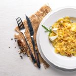 Pasta ai quattro formaggi: la ricetta per farla cremosa e avvolgente 1 In primo piano su un tavolo chiaro c'è un piatto fondo con la pasta ai quattro formaggi. Sulla sinistra delle posate su una tovaglietta in juta e dei grani di pepe nero.