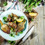 Carciofi alla romana: ricetta e video ricetta di un delizioso contorno sano 1 su tavolo di legno, piatto azzurro con 3 carciofi alla romana, forchetta e coltello accanto, mezzo lime e dietro sale e erbe aromatiche