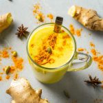 Latte d'oro: perché fa bene, ricetta e video ricetta per preparare il golden milk a casa 1 su tavolo di marmo, tazza trasparente con latte d'oro e attorno polvere di curcuma e zenzero e anice stellato